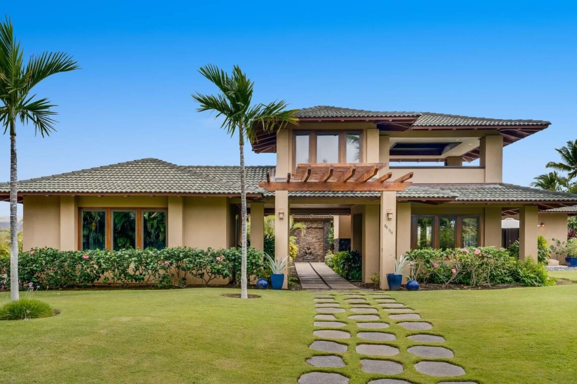 A front view of a luxury two-story villa with a manicured green lawn, palm trees, stone paver paths, a wooden pergola accent over the entryway, and a tiled roof under a clear blue sky.