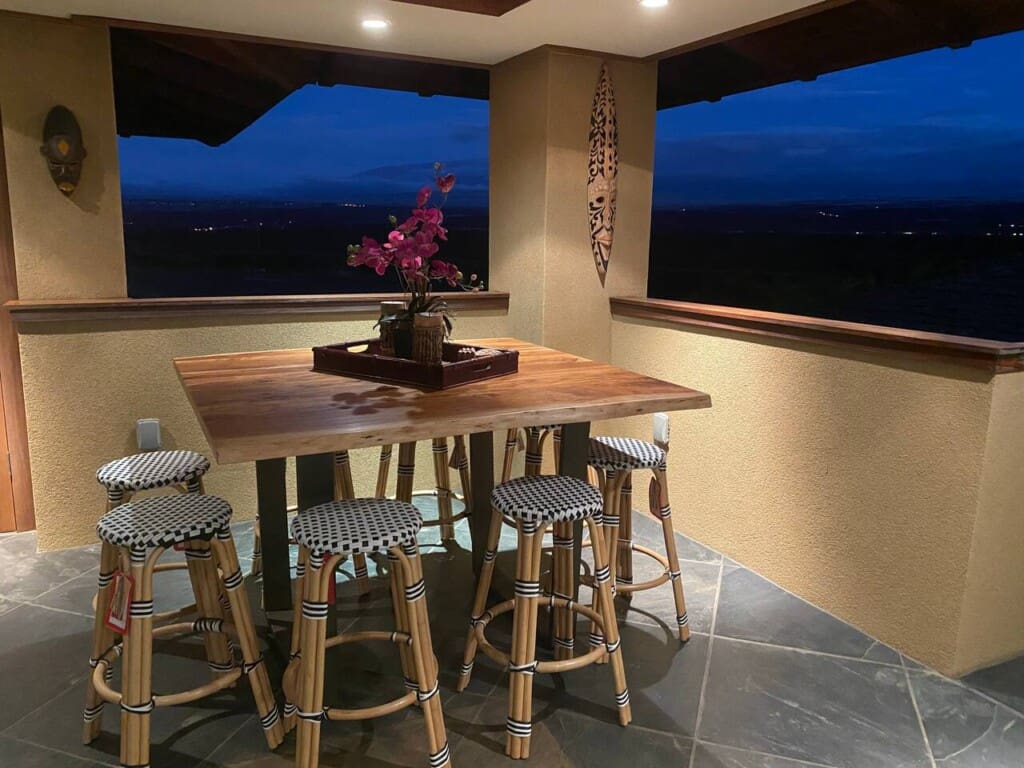 An evening view of an upstairs covered patio featuring a tall wooden bar table, woven bistro-style barstools, tile flooring, and open windows looking out to a dark landscape.
