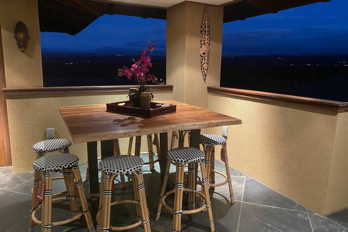 An evening view of an upstairs covered patio featuring a tall wooden bar table, woven bistro-style barstools, tile flooring, and open windows looking out to a dark landscape.