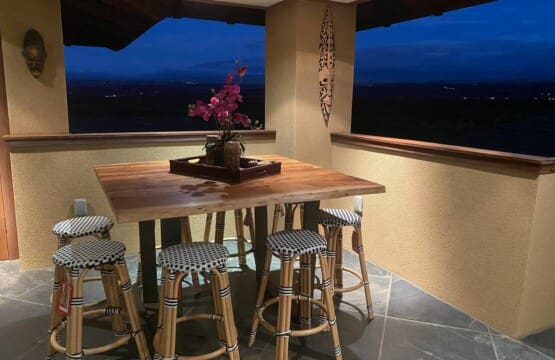 An evening view of an upstairs covered patio featuring a tall wooden bar table, woven bistro-style barstools, tile flooring, and open windows looking out to a dark landscape.