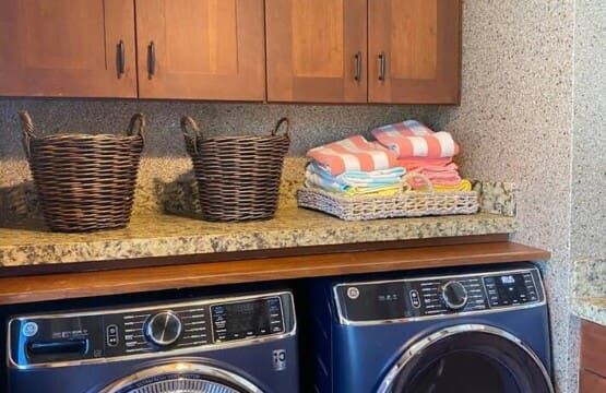 A modern laundry room featuring a blue front-loading washer and dryer set, a stone countertop, woven baskets, and wooden upper cabinets.