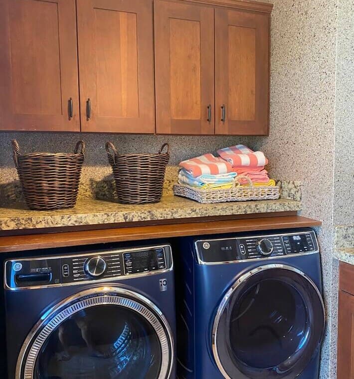 A modern laundry room featuring a blue front-loading washer and dryer set, a stone countertop, woven baskets, and wooden upper cabinets.