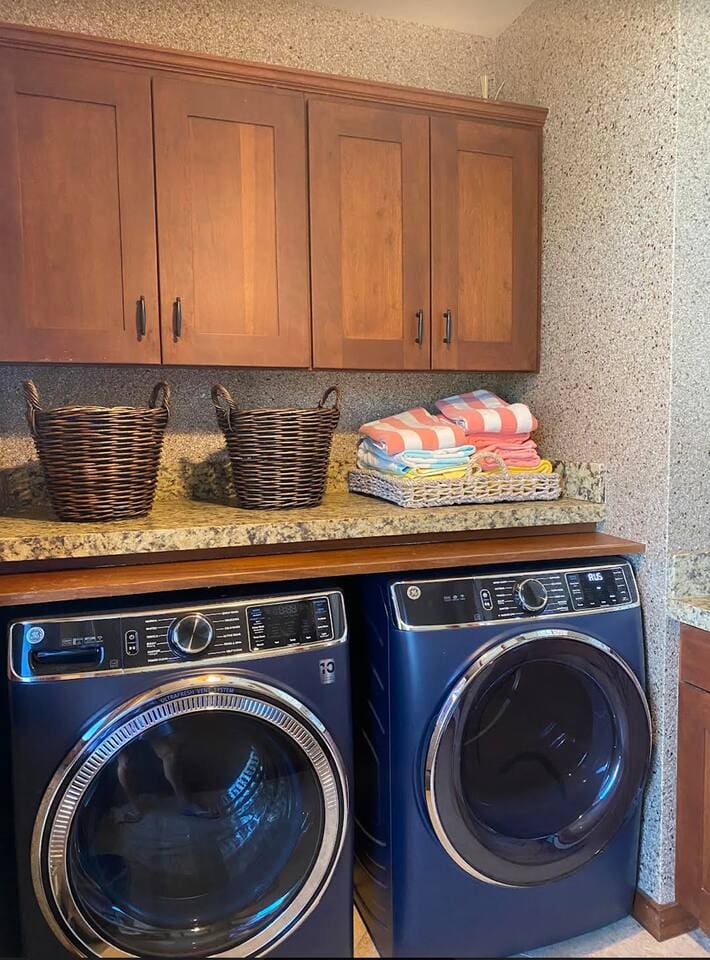 A modern laundry room featuring a blue front-loading washer and dryer set, a stone countertop, woven baskets, and wooden upper cabinets.