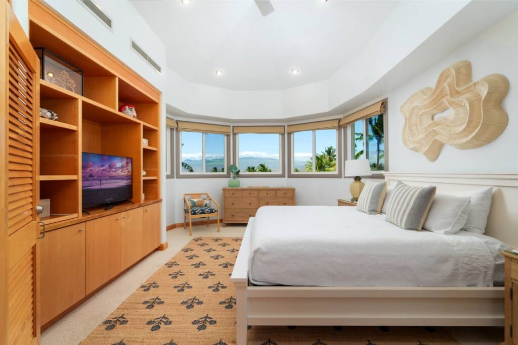Bright bedroom at a Mauna Lani luxury vacation rental featuring a large bed, wooden built-in shelving with a TV, and windows showing distant mountains.