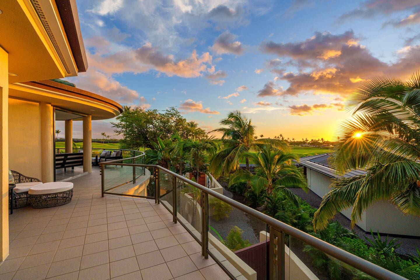 An upper-level outdoor lanai with glass railings overlooking palm trees and a vibrant tropical sunset.