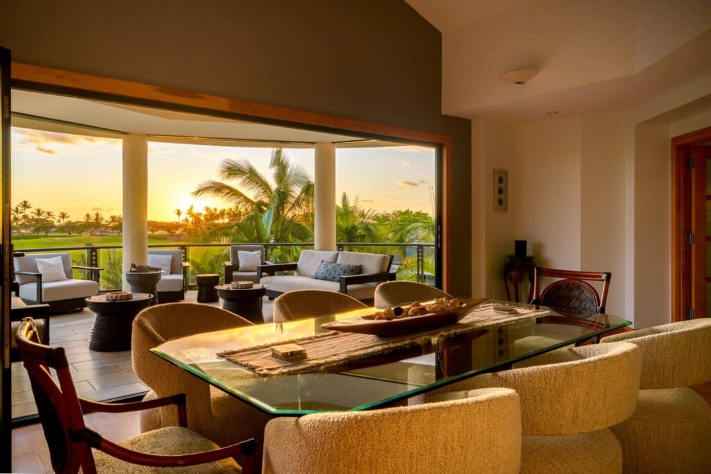 Alt Text An indoor dining area with a glass table looking out to a covered lanai at sunset with palm trees.