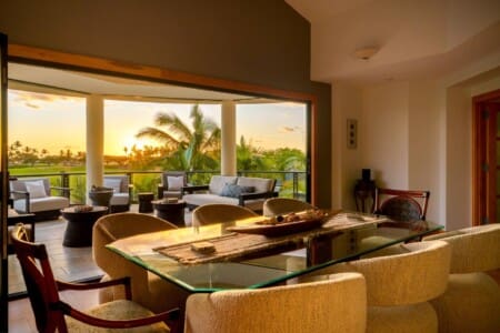Alt Text An indoor dining area with a glass table looking out to a covered lanai at sunset with palm trees.