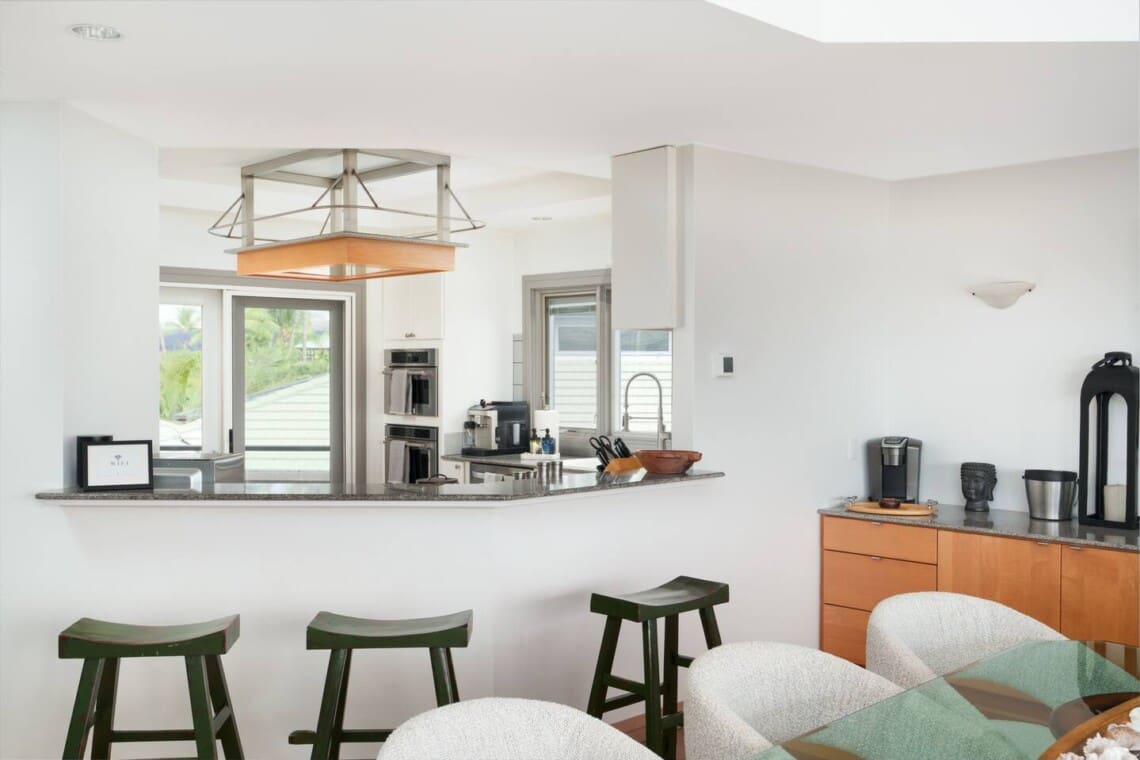 A kitchen breakfast bar with green wooden stools adjacent to an indoor dining table with upholstered chairs.