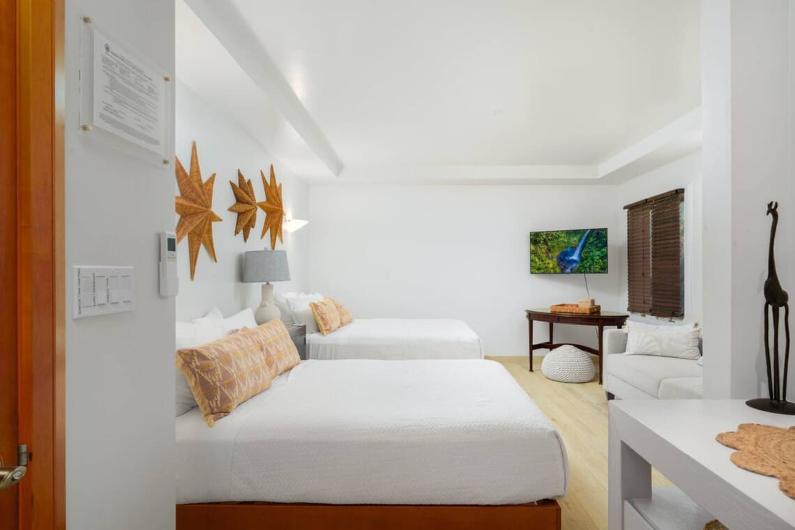 Bright guest bedroom at a Mauna Lani vacation home featuring two beds, a wall-mounted TV, and woven star decor.