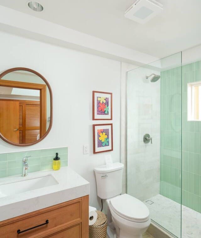 Bright modern bathroom featuring a wooden vanity, round mirror, white toilet, and a glass walk-in shower with mint green tiles.