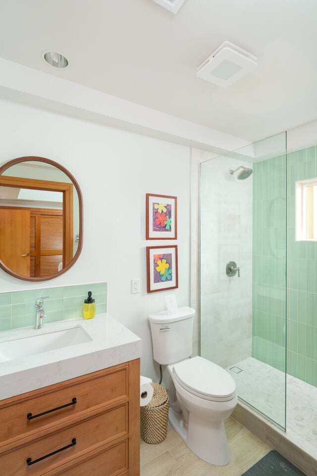 Bright modern bathroom featuring a wooden vanity, round mirror, white toilet, and a glass walk-in shower with mint green tiles.