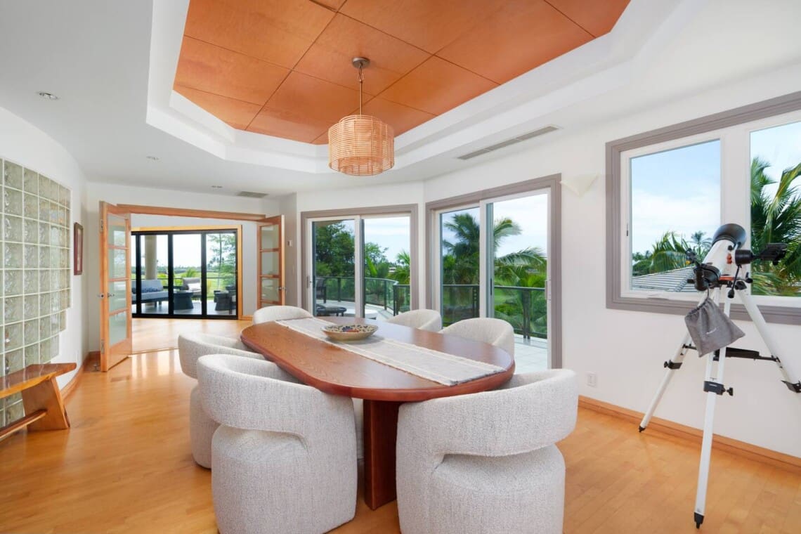 A bright secondary dining or meeting room with an oval wooden table, upholstered chairs, and a telescope near a window.