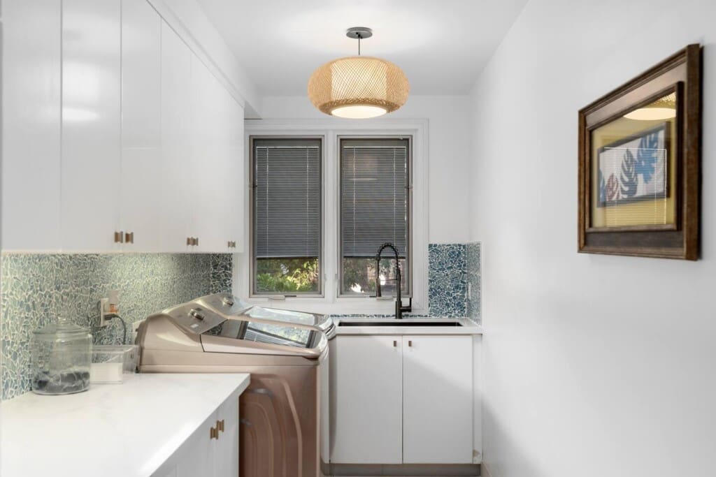 A bright laundry room with white upper and lower cabinets, a sink, a woven light fixture, and a washing machine.