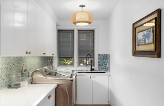 A bright laundry room with white upper and lower cabinets, a sink, a woven light fixture, and a washing machine.