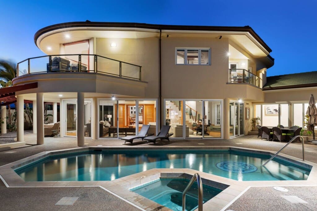 Alt Text Rear exterior view of a two-story luxury villa illuminated at dusk, featuring a private swimming pool and hot tub surrounded by a spacious lounge patio.