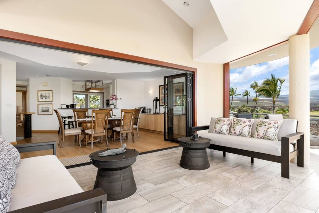 A bright, covered outdoor lanai with modern sofas and unique dark metal coffee tables that opens seamlessly through large sliding glass pocket doors into an indoor dining area with a wooden table and chairs.