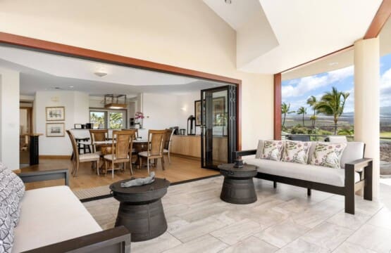A bright, covered outdoor lanai with modern sofas and unique dark metal coffee tables that opens seamlessly through large sliding glass pocket doors into an indoor dining area with a wooden table and chairs.