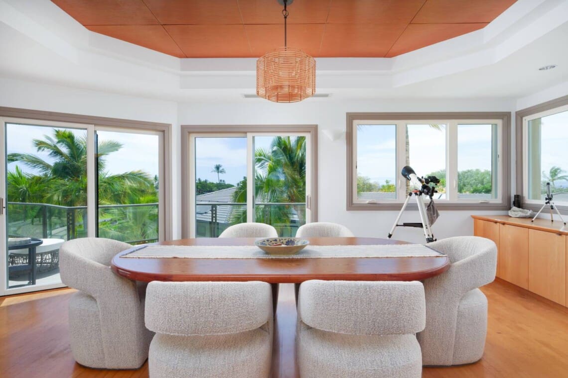 An upper-level dining space featuring a wooden table, upholstered chairs, a wicker chandelier, and a telescope near large windows.