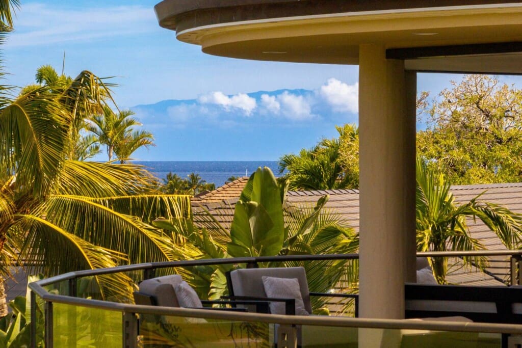 An upper-level outdoor balcony with comfortable lounge chairs framed by lush palm trees, offering distant views of the ocean and mountains.