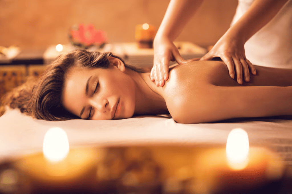 A woman lying down with her eyes closed receiving a relaxing back massage from a professional therapist, with warm candles glowing in the blurred foreground.